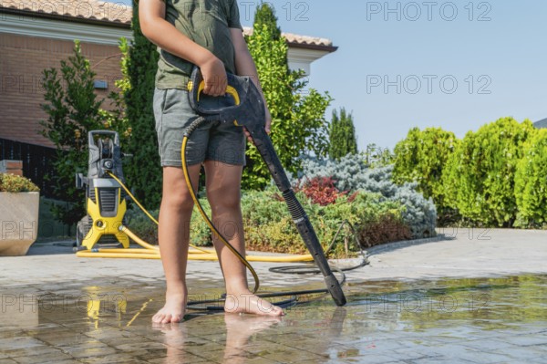 A boy in shorts uses a pressure washer to clean a garden. The washer is in the background, and the scene is surrounded by greenery under a clear blue sky