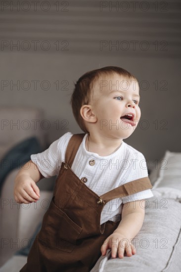 A cheerful toddler in brown overalls and a white shirt plays joyfully on a soft sofa. The playful expression on the child face captures innocence and happiness