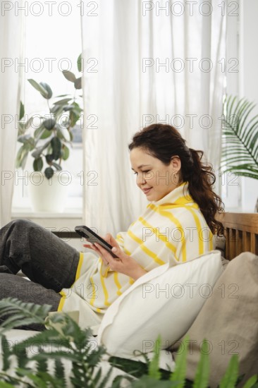 A woman lounges on a sofa, interacting with her smartphone. Surrounded by plush pillows and indoor plants, she enjoys a peaceful moment in a cozy, sunlit room