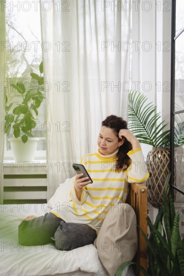 A woman in a striped yellow sweater relaxes on a bed using her smartphone. The room has a cozy ambiance with greenery and natural light pouring through sheer curtains
