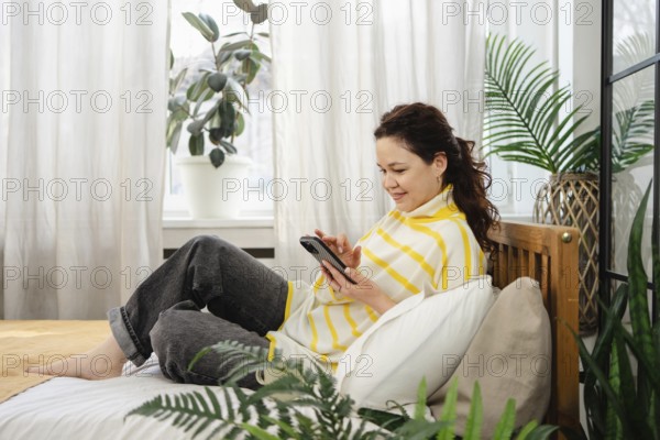 A woman sits comfortably on a bed, wearing a striped top, using her phone. The room is filled with natural light and surrounded by lush green plants, exuding a sense of calm and relaxation