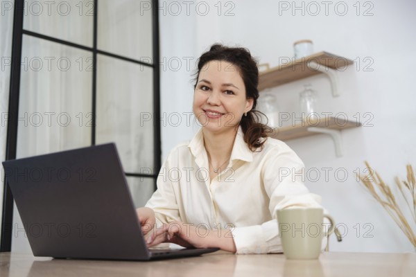 A professional woman, smiling and working on her laptop at a home office desk. Shelves and plants in the background create a warm and productive atmosphere