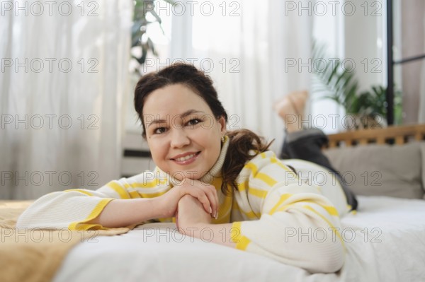 A woman in a yellow striped sweater smiles while lying on a comfortable bed, surrounded by soft pillows and bright natural light. The scene conveys warmth and relaxation