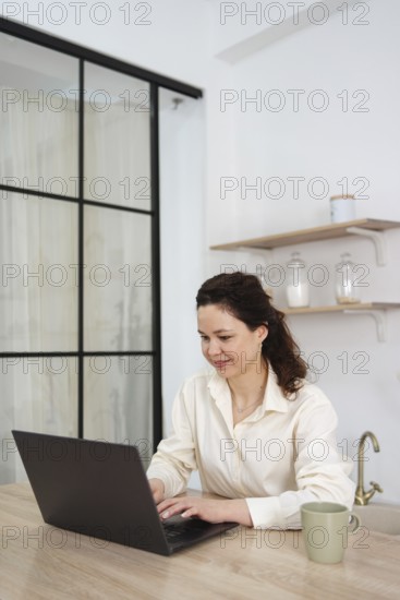 A woman works from home, sitting at a table with a laptop. She is focused and looks at the screen, suggesting remote work or freelance activity in a calm environment