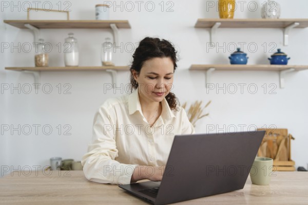 A woman sits at a table, focused on her laptop. Shelves with jars and decor create a cozy, organized atmosphere, representing remote work or study from home