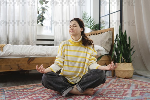 A woman sits cross legged on the floor of a cozy, sunlight filled bedroom at home, meditating in a yoga pose. She is calm and relaxed, wearing a striped yellow sweater