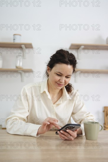 A woman in a cream shirt sits at a wooden table at home using her smartphone, next to a green coffee cup, with shelves adorned with jars in the background