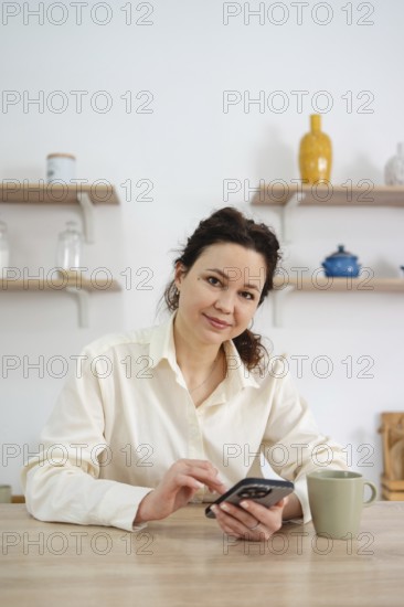 A woman in a white blouse sits at a wooden table in her kitchen, using a smartphone. A mug is beside her, while shelves with colorful decor are in the background