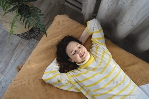 A woman lies on a brown blanket, smiling and relaxed, wearing a white and yellow striped sweater. A plant beside her adds a touch of nature to the calming scene