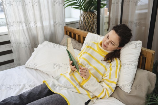 A woman in a yellow striped sweater is relaxing on a bed while reading a book. Soft natural lighting filters through curtains, creating a serene and inviting atmosphere