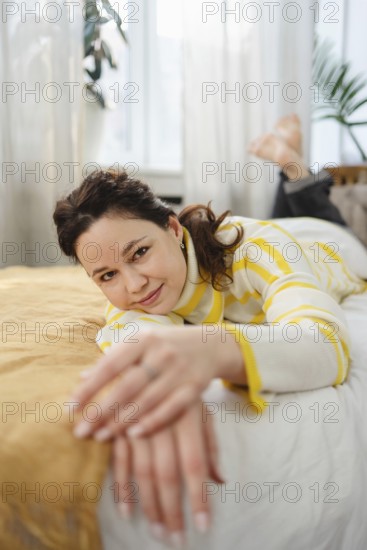 A woman in a striped sweater enjoys a relaxing moment at home. She lies on a bed in a sunlit room, exuding warmth and comfort. Soft natural light enhances the cozy atmosphere