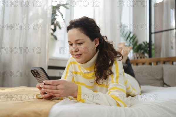 Smiling woman wearing a striped sweater lays on her bed and uses her smartphone. The bright and cozy room is filled with natural light and houseplants