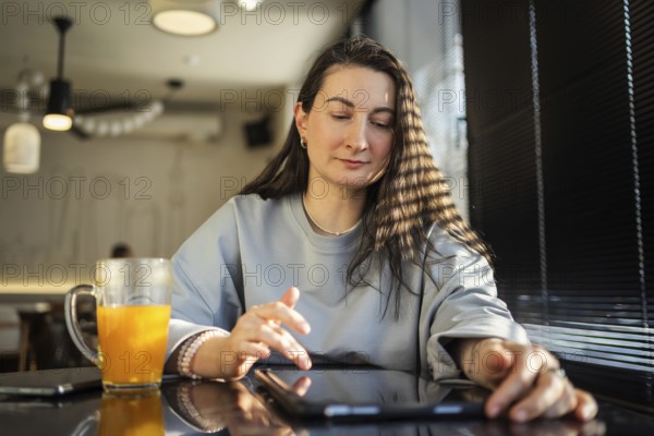 A serene middle-aged woman interacts with a tablet at a sunlit cafe table, amidst a relaxed setting with a drink by her side