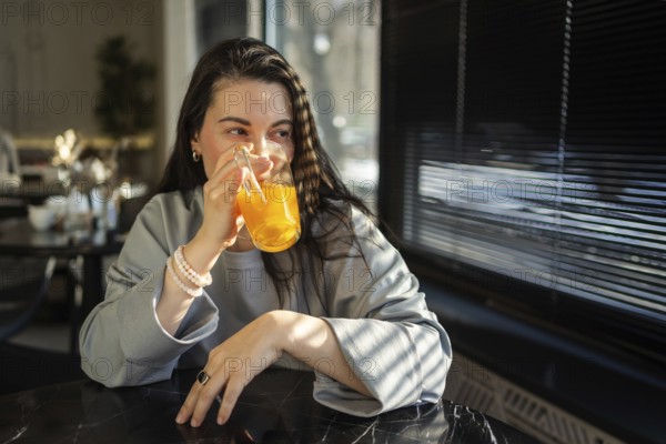 A mid-aged woman sips an orange drink in a brightly lit cafe, her expression serene and content as sunlight streams through the blinds