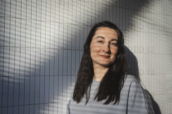A middle-aged woman smiles gently while standing against a geometrically patterned tile wall, with sunlight casting diagonal shadows