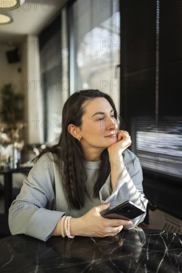 A middle-aged woman sits peacefully in a cafe, gazing outside while holding a smartphone in her hand. The sunlight highlights her thoughtful expression
