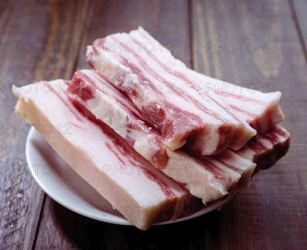 Slices of raw pork belly are neatly arranged on a white plate set against a rustic wooden surface. The marbled fat and rich red meat highlight its freshness