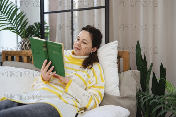 A woman wearing a striped sweater lies on a comfortable bed, deeply engrossed in reading a green book. The scene is enhanced by lush green plants, creating a serene atmosphere