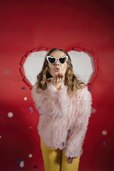 A charming woman with long hair wearing heart-shaped sunglasses and a frilly pink blouse sends a kiss against a festive red background, celebrating Valentine's day
