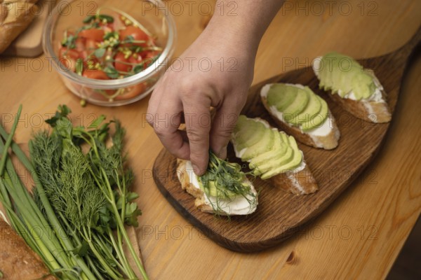 A hand garnishes avocado toast with fresh herbs on a wooden board. Nearby are a salad with tomatoes and a bunch of herbs on a wooden table, creating a healthy meal setting