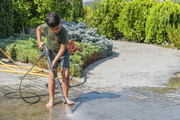 A child is using a pressure washer to clean a stone patio for the summer. He is barefoot, wearing a green shirt, surrounded by lush greenery. Water splashes from the powerful stream