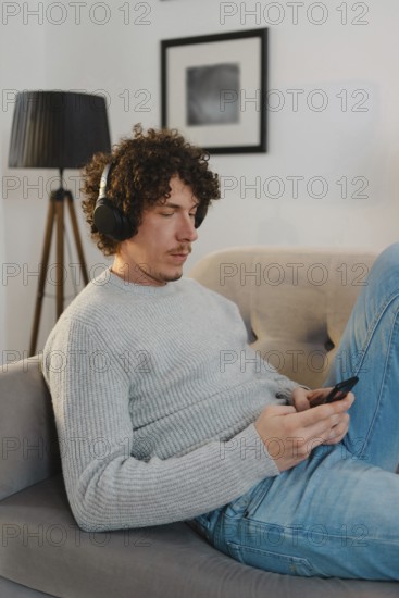 A young man with curly hair relaxes on a sofa, wearing a gray sweater, blue jeans, and headphones. He is focused on his phone, creating a calm atmosphere