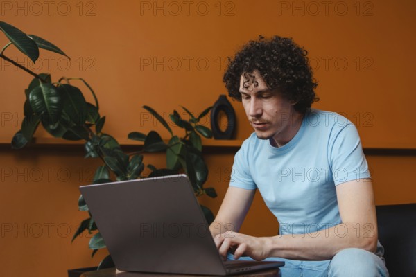A young man with curly hair focused on his laptop in a modern room with orange walls and green plants, wearing a blue t-shirt, conveying a creative work atmosphere