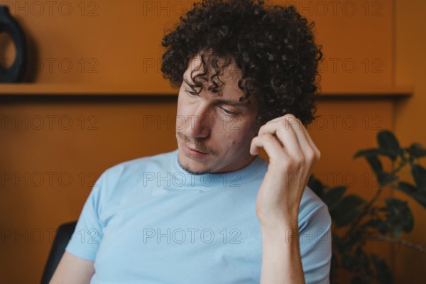 A young man with curly hair in a light blue shirt, sitting thoughtfully in a room with an orange background and green plants. The scene conveys a casual, introspective mood