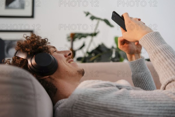 A young man with curly hair enjoying leisure time on a sofa, wearing headphones and using a smartphone. The relaxed atmosphere showcases modern technology use