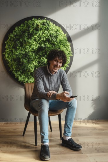 A young man with curly hair, sitting in a chair next to a vibrant green wall, embodies themes of ecology and sustainable development in a modern setting