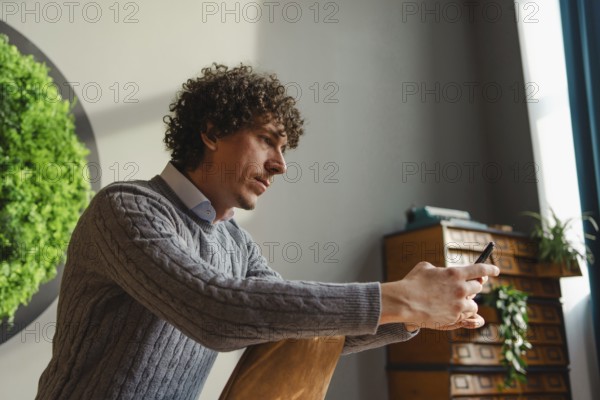 A young man with curly hair uses a smartphone, sitting beside a vibrant green wall. This scene highlights themes of ecology and sustainable development in a modern setting