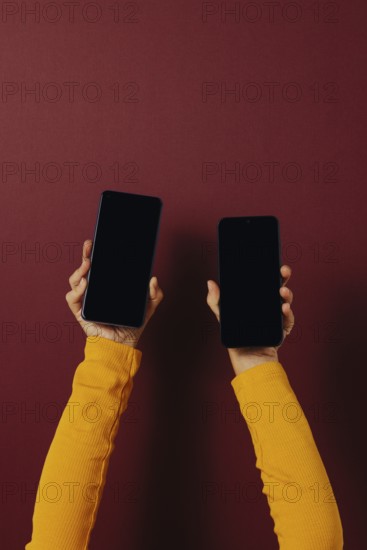 Side view of unrecognizable person in casual clothes holding smartphones with black screen while standing against dark red background
