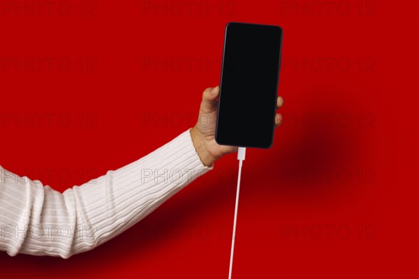 Side view of unrecognizable person in casual clothes holding smartphone with black screen while standing against red background