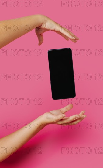 Crop anonymous person in casual clothes showing modern smartphone with empty screen while floating between hands on pink background