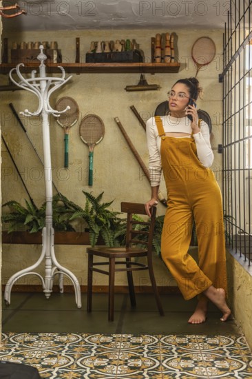 A young woman engages in teleworking, standing while using a smartphone, dressed in bold mustard overalls, surrounded by eclectic decor and homely elements