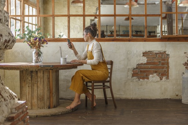 A young woman uses a smartphone and laptop while sitting at a round wooden table in a rustic cafe, displaying elements of teleworking in an informal environment