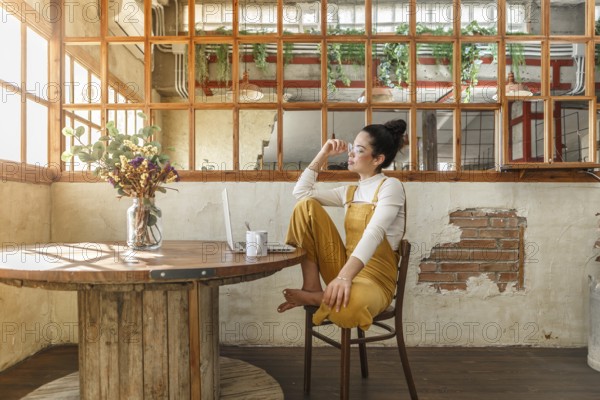 A young woman enjoys a coffee break in a sunlit rustic loft, with a laptop nearby, illustrating a relaxed teleworking environment