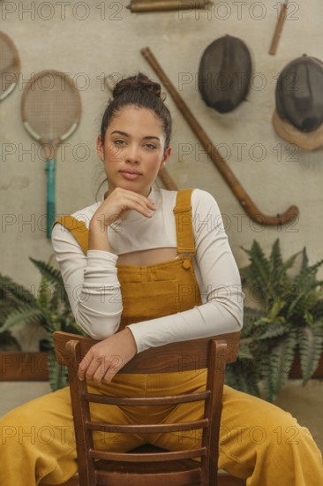A young woman leans on a vintage wooden chair, dressed in a mustard yellow jumpsuit with a playful air, surrounded by rustic decor and sporting equipment on the walls