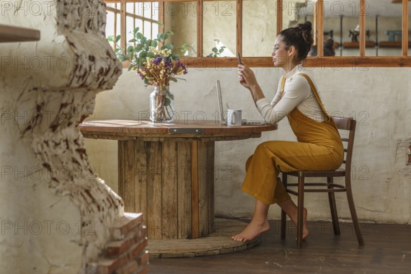 A woman uses a smartphone and laptop at a wooden table in a rustic setting, emphasizing the blend of modern telework with traditional decor