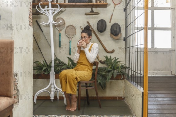 A woman in a yellow jumpsuit sips coffee in her eclectic teleworking space, surrounded by vintage sports equipment and plants