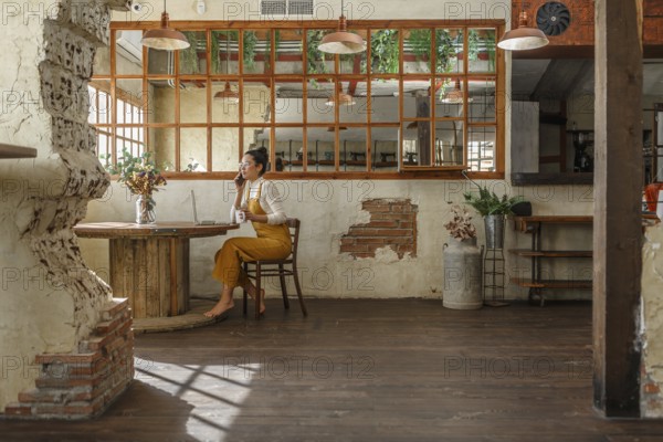 A woman teleworking in a vintage style cafe, using a laptop and smartphone as she engages in a phone conversation