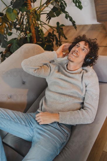 Young man with curly hair relaxes on a modern gray couch, wearing headphones and enjoying music. He has a cozy sweater, blue jeans, and a content expression