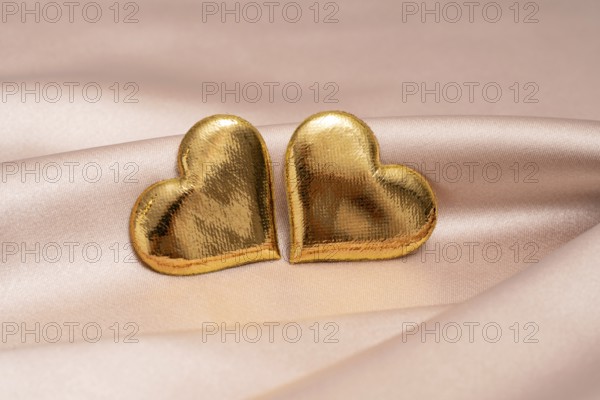 Two golden heart-shaped objects rest on a smooth pink satin surface, symbolizing love and Valentine's Day in a soft, romantic setting