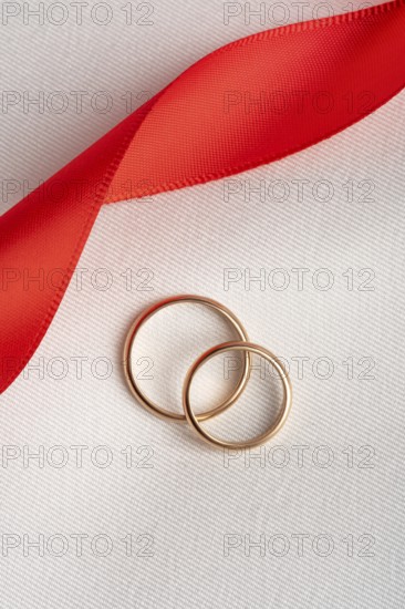 Top view of two golden wedding rings lying on a textured white fabric, intertwined with a vibrant red ribbon, symbolizing romance and eternal love, ideal for St. Valentine's Day themes and wedding concepts