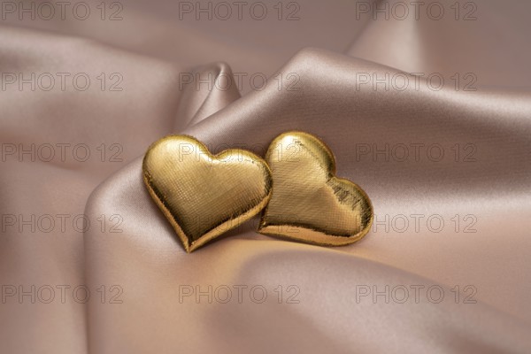 Two golden heart-shaped objects rest gently on a smooth, silky fabric, symbolizing love and commitment, perfect for St. Valentine's Day and wedding themes