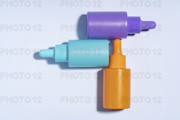 Top view of three vibrant serum bottles in purple, turquoise, and orange colors, designed with droppers, displayed on a clean, light blue background