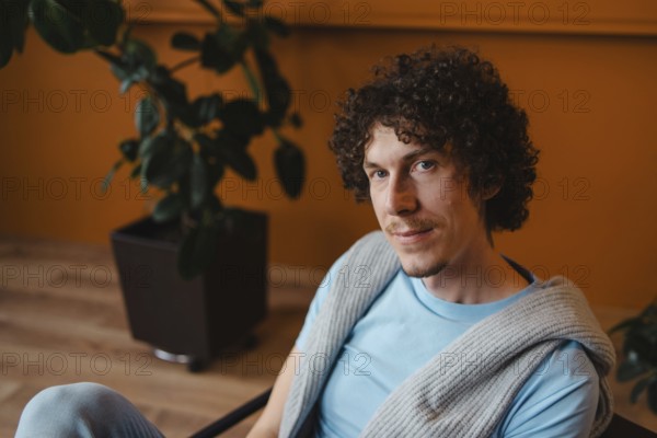 Young man with curly hair in a cozy setting, wearing a blue shirt and a knit sweater. Warm-toned room with plants, creating a relaxed and natural atmosphere