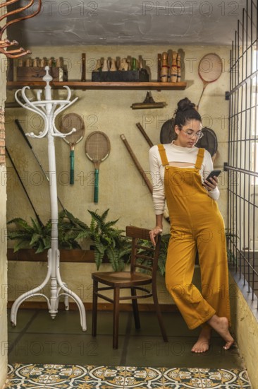 A young woman dressed in fashionable yellow overalls and glasses uses a smartphone while standing in a rustic indoor area with vintage sports equipment