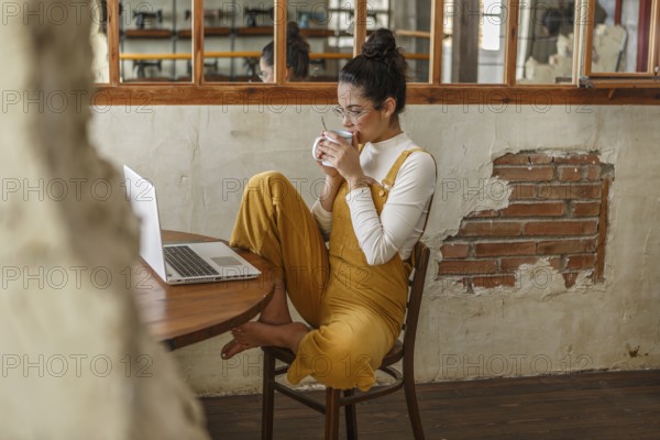 A young woman teleworking in a rustic home setting, sipping tea while using her laptop