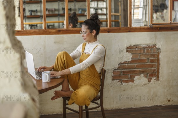 A young woman in a stylishly rustic space focuses on her laptop screen while teleworking, her coffee mug nearby inviting a relaxed work atmosphere